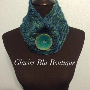 May include: A teal blue and green knitted neck warmer with a large, round, teal blue button in the center. The text "Glacier Blu Boutique" is printed below the neck warmer.