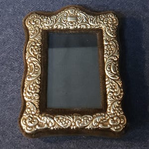 May include: An antique silver-coloured picture frame with a dark brown velvet backing. The frame has detailed floral and scrollwork patterns. The rectangular frame has a curved top, designed to hold a small photograph or artwork.
