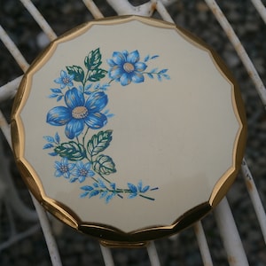 May include: A gold compact with a white background and a blue floral design. The flowers are stylized and have a blue center with white petals.