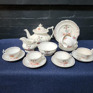 May include: A vintage tea set comprising a white teapot, creamer, sugar bowl, plates, and teacups with saucers. The set features a floral pattern with red, yellow, and green accents. The teacups and saucers are arranged on a blue surface.