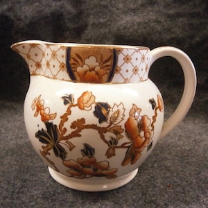 May include: Cream-colored ceramic pitcher with a rounded body and a curved handle. The pitcher is decorated with orange, navy, and brown floral patterns and a geometric design around the rim. A classic piece for serving.