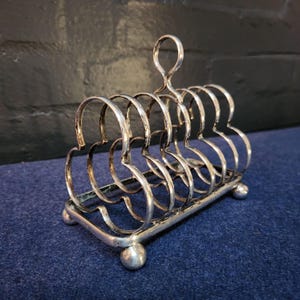 May include: Silver plated toast rack with a decorative design. The rack has six slots and sits on a rectangular base with rounded feet.