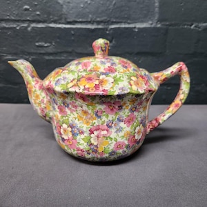 May include: A floral-patterned teapot with a matching lid. The teapot features a handle and spout, and is covered in a colorful pattern of pink, yellow, blue, and green flowers on a white background. The teapot is made of ceramic.