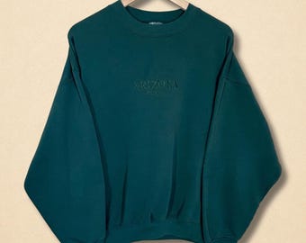 May include: A dark teal sweatshirt with the word "ARIZONA USA" embroidered on the front. The crew neck sweatshirt is hanging on a wooden hanger. The garment has a relaxed fit.