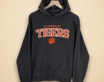 May include: Black hooded sweatshirt with the text "CLEMSON TIGERS" in orange and white lettering. Below the text is an orange paw print. The sweatshirt has a front pocket and a drawstring hood.