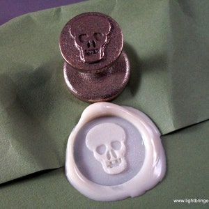 May include: A close-up of a brass wax seal stamp with a skull design. The stamp is used to create a wax seal with the same skull design.