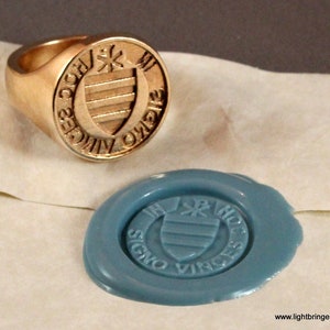 May include: A gold signet ring with a crest featuring a shield with horizontal stripes and a star above it. The ring has the inscription "SIGNO VINCES" around the crest. A blue wax seal with the same crest is shown next to the ring.