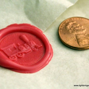 May include: A red wax seal with a train design. The seal is on a white surface next to a copper penny.