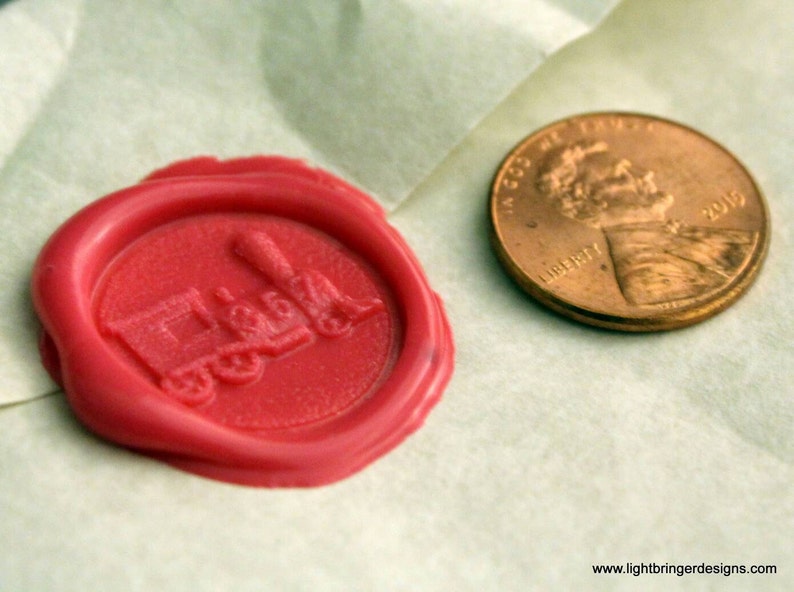May include: A red wax seal with a train design. The seal is on a white surface next to a copper penny.