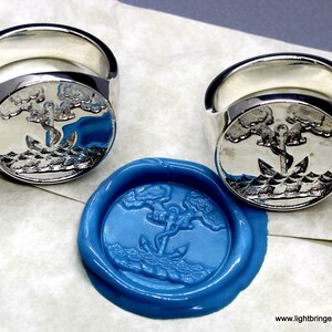 May include: Two silver signet rings with an anchor and two trees design. The rings are shown next to a blue wax seal with the same design.