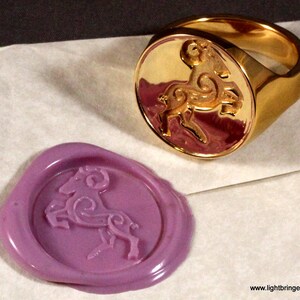 May include: A gold signet ring with a detailed design of a ram or goat. The ring is shown next to a purple wax seal with the same design.