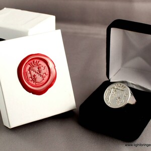 May include: A silver signet ring with a crest design in a black velvet ring box. The ring is displayed on a white background with a red wax seal.