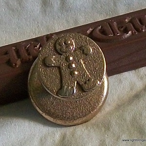 May include: A gold-toned metal gingerbread man cookie cutter with a detailed design. The cutter is round and has a raised gingerbread man figure in the center.