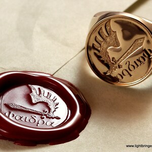 May include: A gold signet ring with a detailed engraving of a winged creature holding a sword. The inscription "pax" is visible on the ring. A red wax seal with the same design is also shown.