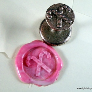 May include: A pink wax seal with a design of two crossed candy canes. The seal is made with a metal stamp that has a similar design.