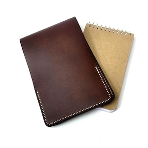 Puede incluir: Una funda de cuaderno de cuero marrón oscuro con costuras blancas y un bloc de notas con espiral. La funda de cuero es rectangular y ligeramente más grande que el bloc de notas, que tiene una cubierta de papel kraft.