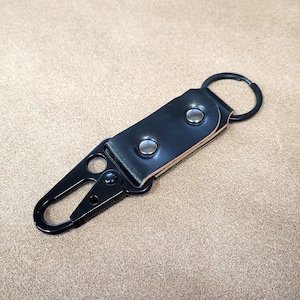 Puede incluir: Llavero de cuero negro con un mosquetón de metal negro y un llavero plateado. La correa de cuero está asegurada con dos remaches plateados. El llavero está diseñado para sujetar llaves u otros objetos pequeños.