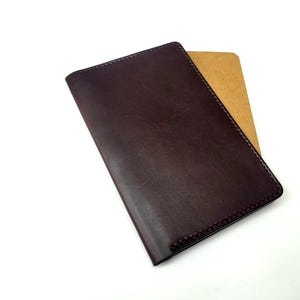 Puede incluir: Una cartera de cuero marrón oscuro con un borde cosido. Un cuaderno de color beige es parcialmente visible en el interior. La cartera rectangular parece estar hecha de cuero de alta calidad.