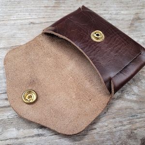 May include: A brown leather wallet with a snap closure and a light brown suede interior. The wallet has two brass snaps.