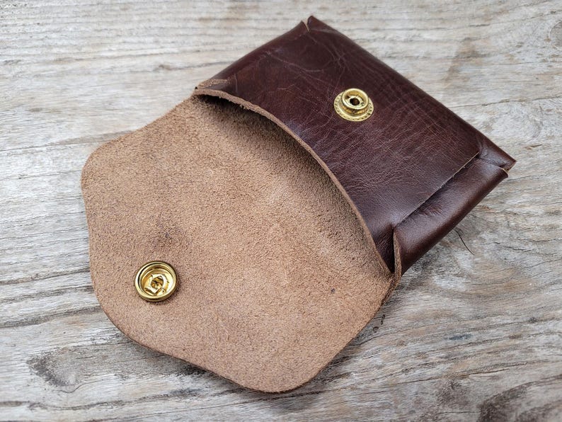 May include: A brown leather wallet with a snap closure and a light brown suede interior. The wallet has two brass snaps.