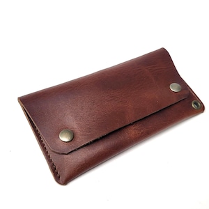 May include: A rectangular brown leather wallet featuring a fold-over flap secured by two metallic snap buttons. The wallet has visible stitching along the edges and a small metal eyelet located on the right side of the flap for attachment.