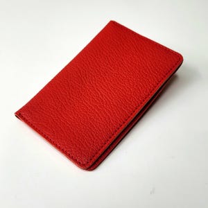 Könnte beinhalten: Ein roter Kartenhalter aus Leder, rechteckig, liegt auf einer weißen Oberfläche. Der Kartenhalter hat eine strukturierte Oberfläche und scheint aus echtem Leder zu sein. Die Kanten sind sauber vernäht.