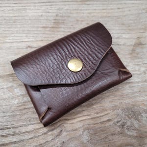 May include: A brown leather wallet with a flap closure and a brass button. The wallet has a textured surface and is folded in half.