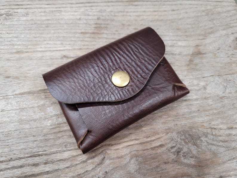May include: A brown leather wallet with a flap closure and a brass button. The wallet has a textured surface and is folded in half.