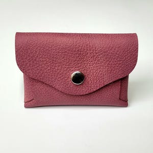 Puede incluir: Una pequeña cartera de cuero burdeos con superficie texturizada. La cartera tiene una solapa curva asegurada por un broche plateado. La cartera rectangular está diseñada para guardar tarjetas o objetos pequeños.