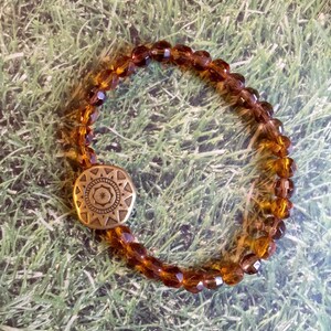 May include: A brown glass beaded bracelet with a gold-toned metal charm featuring a geometric design.