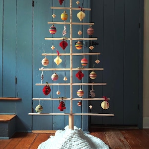 May include: A wooden Christmas tree with horizontal branches, adorned with colorful ornaments of various shapes and sizes. The tree stands on a white knitted base, set against a blue wall and wooden floor.