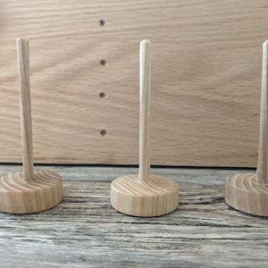 May include: Three wooden thread spool holders. Each holder has a round, grooved base and a tall, slender, cylindrical peg. The wood has a light, natural color. The holders are arranged in a row against a wooden background.