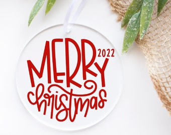 May include: A clear, round Christmas ornament with the words "MERRY Christmas 2022" in red script. The ornament hangs from a white ribbon, with greenery and berries in the background. A festive holiday decoration.