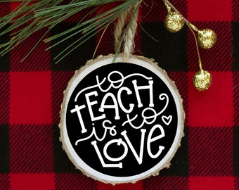 May include: A round wooden ornament with a black background and white lettering that reads "to teach is to love". The ornament is hanging from a piece of twine and is sitting on a red and black buffalo plaid fabric.