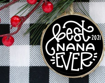 May include: A black and white round wooden ornament with the text "Best Nana Ever" in white cursive lettering and the year "2021" in white text. The ornament is sitting on a black and white checkered surface.