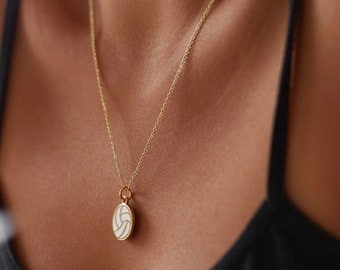 May include: A gold-colored necklace with a pendant shaped like a volleyball. The pendant is white with black lines, and the chain is delicate. The necklace is worn around the neck.