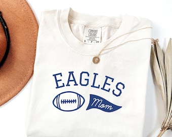 May include: Cream-colored t-shirt with the word "EAGLES" in navy blue block letters, a navy blue football graphic, and the word "Mom" on a navy blue banner. A gold necklace with a pendant is draped on the shirt.