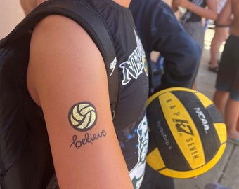 May include: A person's arm with a temporary tattoo of a volleyball with the word "Believe" written underneath. The tattoo is yellow and green. The person is holding a yellow and black volleyball.