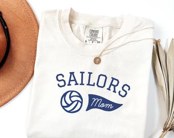 May include: Cream-colored t-shirt with the words "SAILORS Mom" in navy blue, along with a volleyball graphic. A gold necklace with a pendant rests on the shirt. The shirt is a Comfort Colors brand.