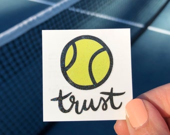 May include: A white sticker with a black outline of a yellow tennis ball and the word "trust" written in black script.