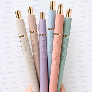 May include: A set of six retractable pens in pastel shades of beige, tan, lavender, light blue, peach, and sage green. Each pen features gold-tone metal accents at the top and center, with the name ASHLEY NICOLE engraved in gold lettering.