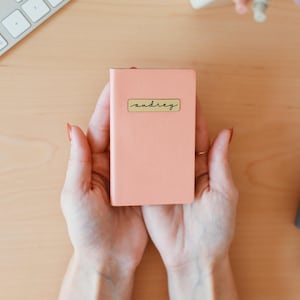 May include: A person's hands holding a small pink notebook with a gold label that says "andrey" on it.