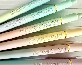 May include: A collection of six pastel-colored pens with gold accents. Each pen is inscribed with a different empowering phrase: "SMASH THE PATRIARCHY," "GIRLS WILL BE GIR POWERFUL," "PEACE. LOVE. FEMINISM.," "NOT HERE TO PLEASE," "I SAID WHAT I SAID," and "RAGE, GRACEFULLY."