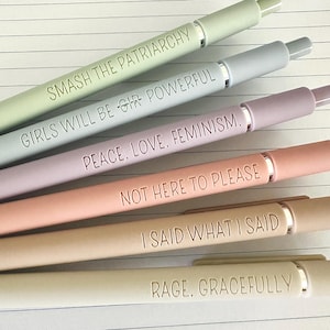 May include: An assortment of pastel-colored pens with silver accents. Each pen is engraved with a different empowering phrase: "SMASH THE PATRIARCHY," "GIRLS WILL BE GIR POWERFUL," "PEACE. LOVE. FEMINISM.," "NOT HERE TO PLEASE," "I SAID WHAT I SAID," and "RAGE, GRACEFULLY."