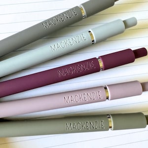 May include: Five pens with the name Mackenzie engraved on them. The pens are in different colors: gray, light blue, burgundy, light pink, and dark gray.