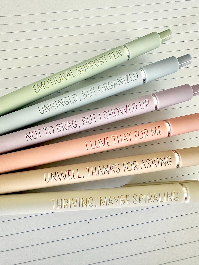Puede incluir: Un conjunto de cinco bol&iacute;grafos de colores pastel con detalles plateados. Cada bol&iacute;grafo tiene una frase grabada: "EMOTIONAL SUPPORT PEN", "UNHINGED, BUT ORGANIZED", "NOT TO BRAG, BUT I SHOWED UP", "I LOVE THAT FOR ME", "UNWELL, THANKS FOR ASKING" y "THRIVING, MAYBE SPIRALING".