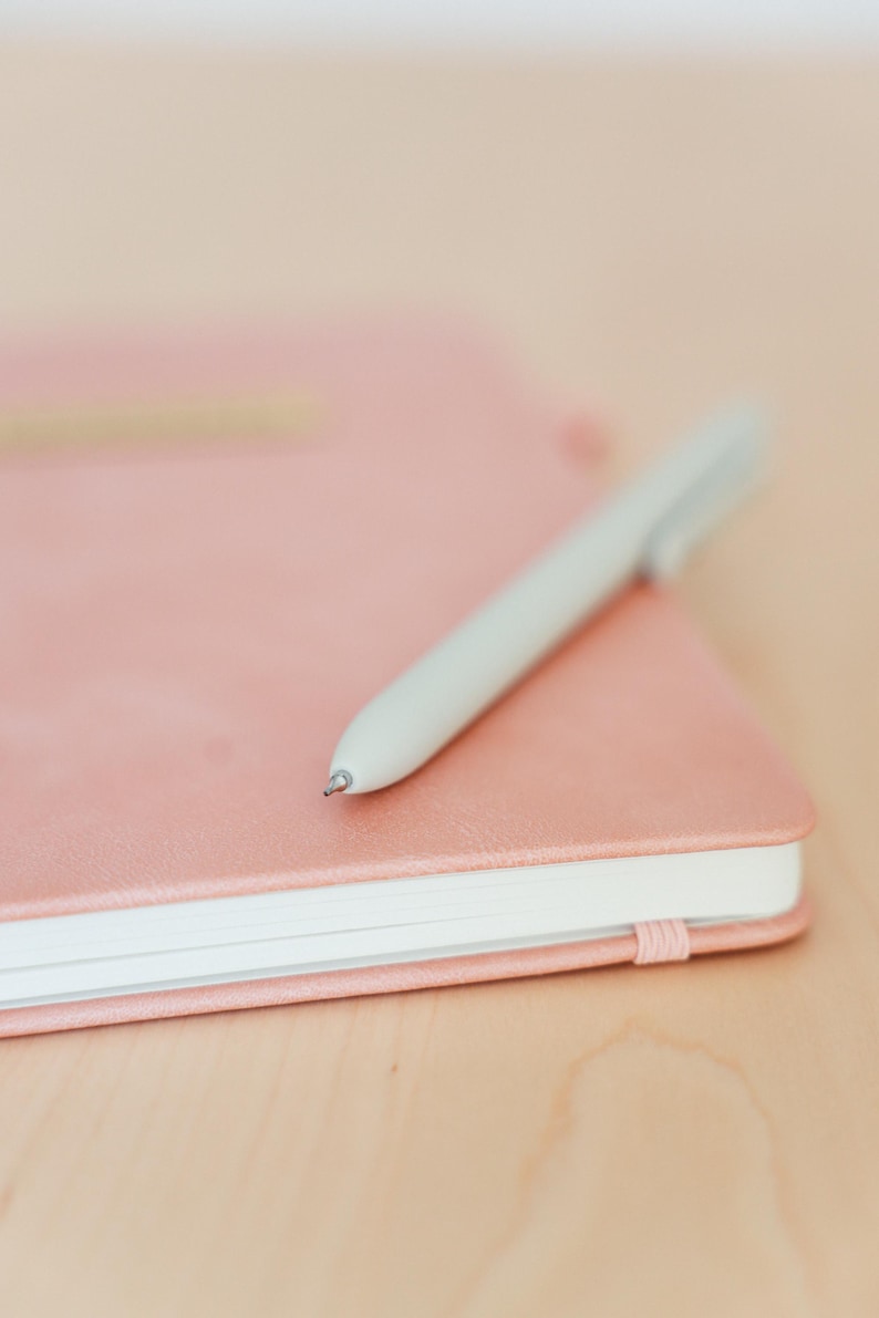 May include: A light pink notebook with a matching light gray pen resting on top. The notebook has a textured cover and a white edge. The pen is angled across the notebook, suggesting it's ready for use. The background is a light wooden surface.