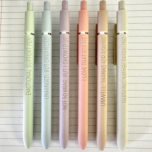 Puede incluir: Seis bol&iacute;grafos de colores pastel con detalles plateados y texto negro. Los bol&iacute;grafos est&aacute;n dispuestos verticalmente y cada uno tiene una frase diferente: "EMOTIONAL SUPPORT PEN", "UNHINGED, BUT ORGANIZED", "NOT TO BRAG, BUT I SHOWED UP", "I LOVE THAT FOR ME", "UNWELL, THANKS FOR ASKING" y "THRIVING, MAYBE SPIRALING".