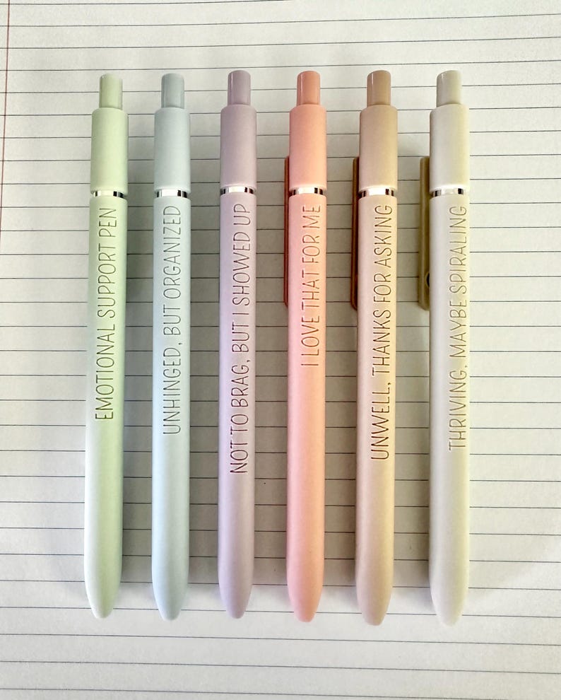 Puede incluir: Seis bol&iacute;grafos de colores pastel con detalles plateados y texto negro. Los bol&iacute;grafos est&aacute;n dispuestos verticalmente y cada uno tiene una frase diferente: "EMOTIONAL SUPPORT PEN", "UNHINGED, BUT ORGANIZED", "NOT TO BRAG, BUT I SHOWED UP", "I LOVE THAT FOR ME", "UNWELL, THANKS FOR ASKING" y "THRIVING, MAYBE SPIRALING".