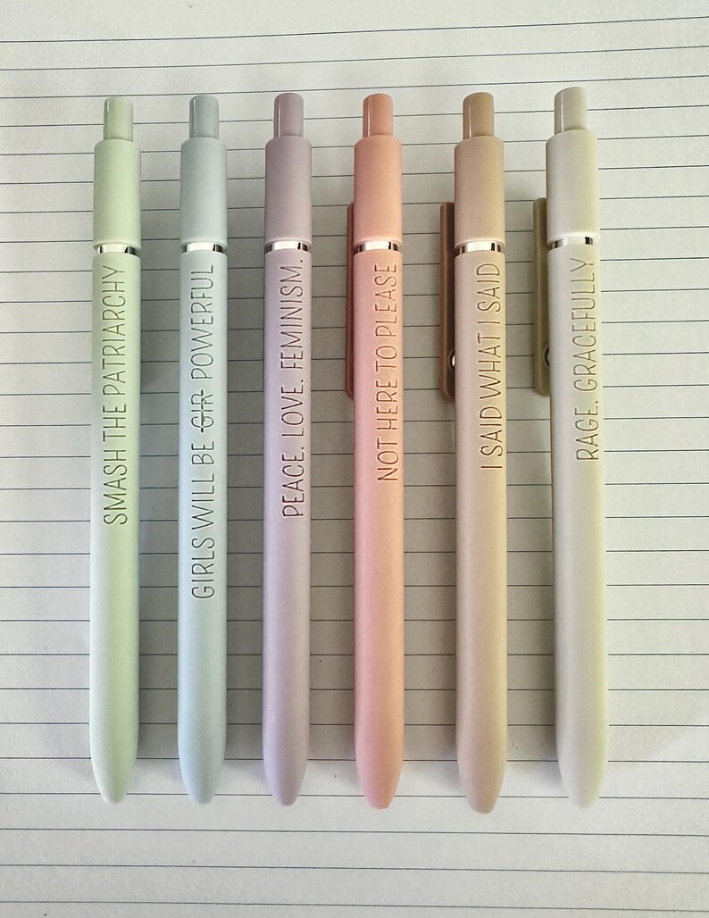 May include: Six pastel-colored pens with gold-colored clips and text. The pens are in shades of green, blue, purple, peach, tan, and white. The text on the pens includes phrases like "SMASH THE PATRIARCHY" and "RAGE, GRACEFULLY."